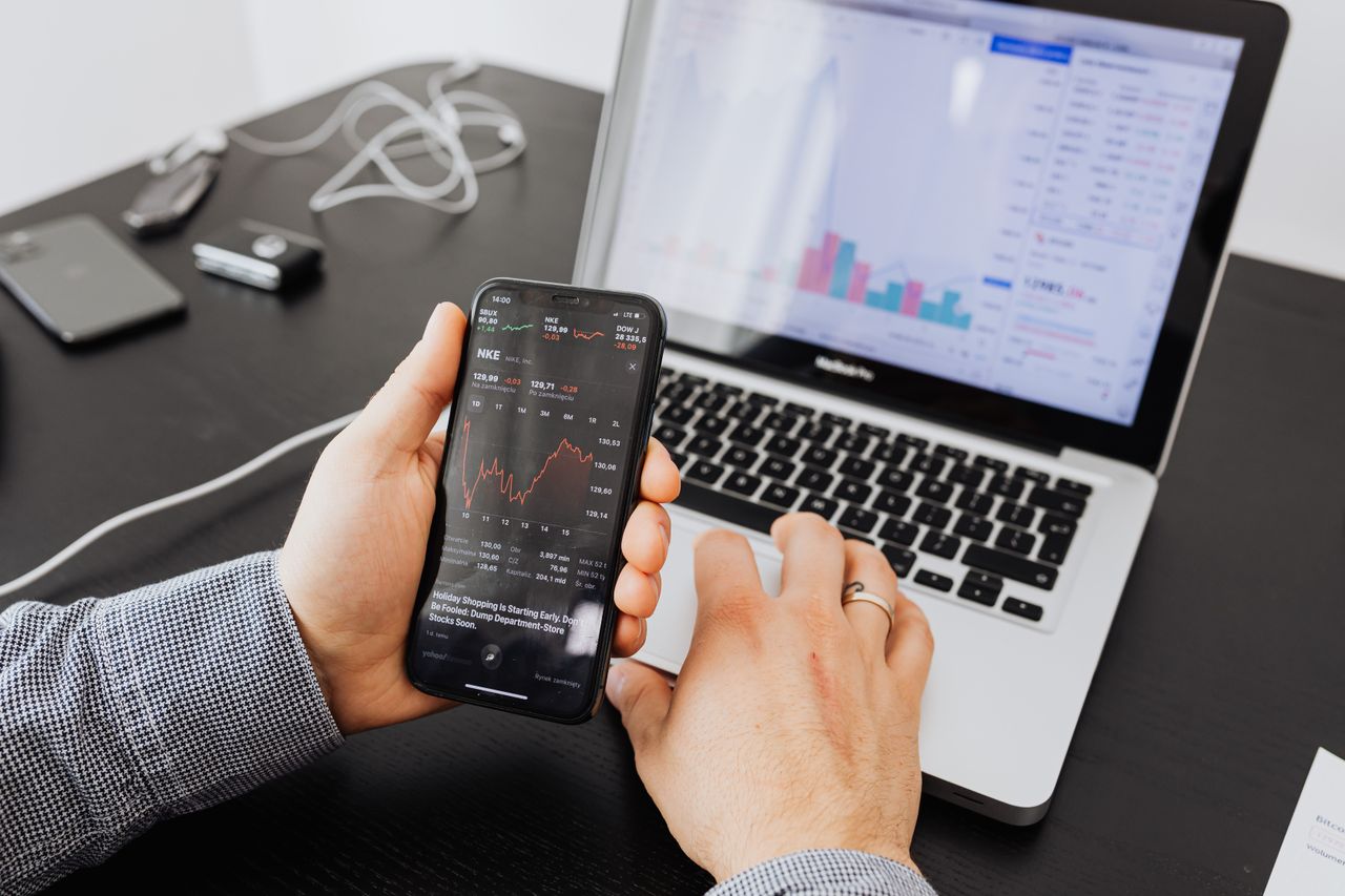 A person reviews stock data on a smartphone and laptop while indoors.
